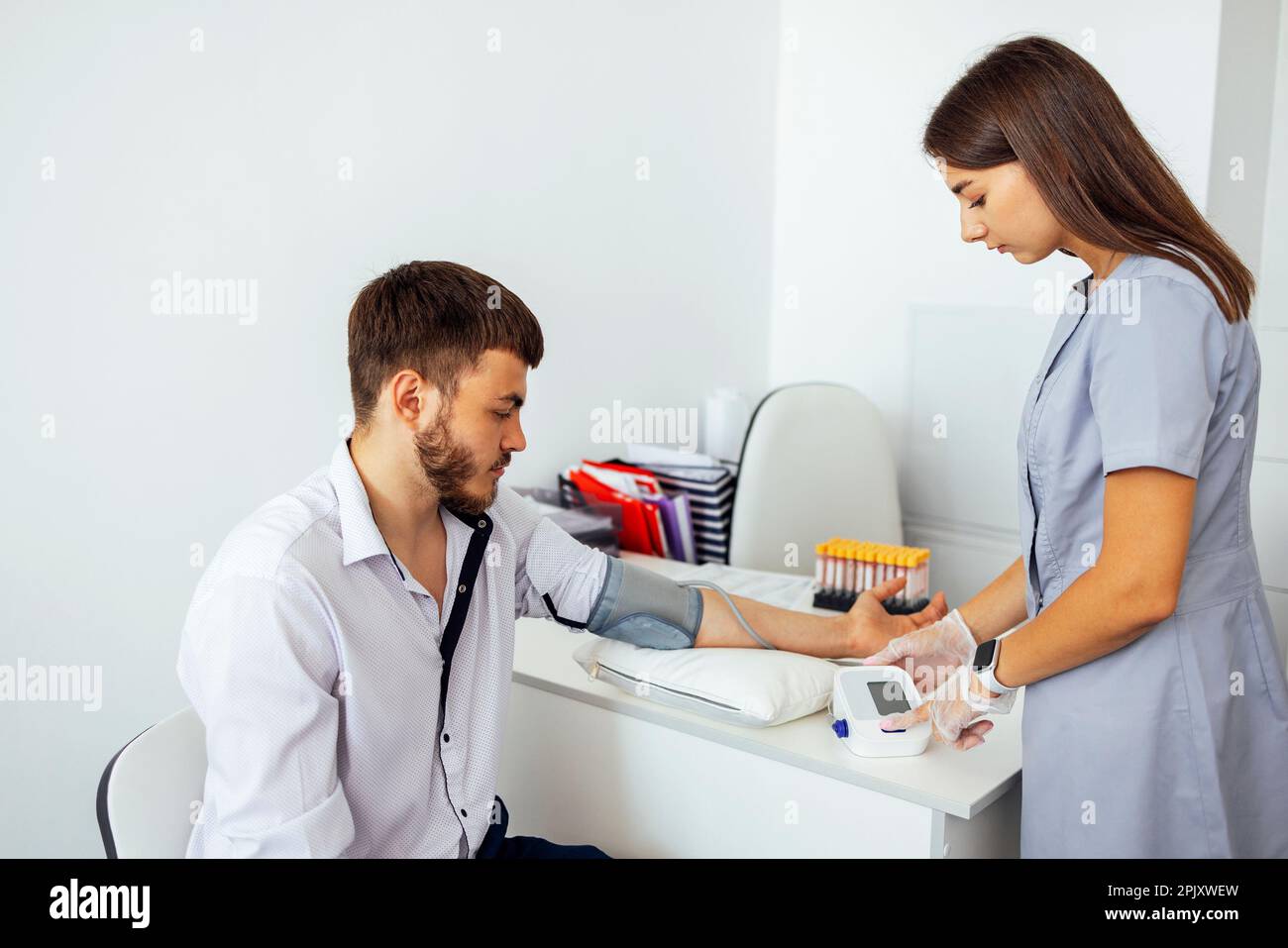 Nurse in medical uniform and transparent gloves checking blood pressure of young man in casual clothes. Brunette woman presses button on tonometer. Te Stock Photo