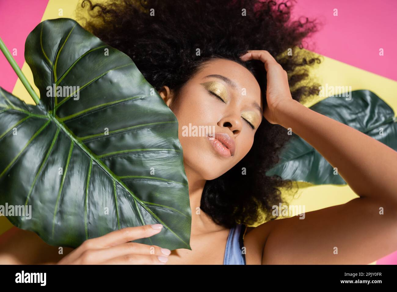 top view of curly african american woman with closed eyes getting tan near green palm leaves on pink Stock Photo