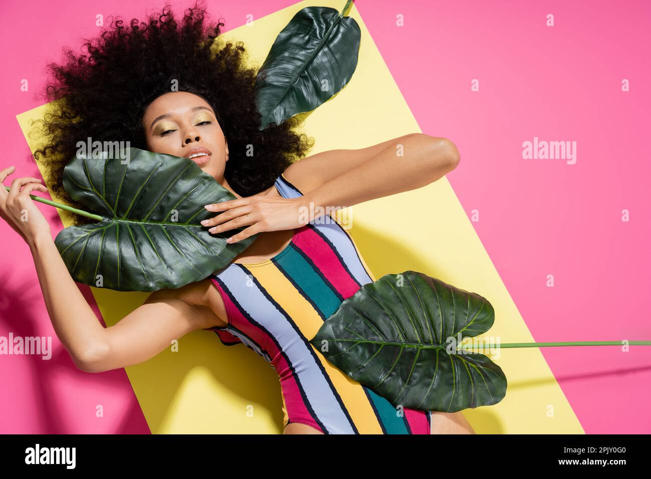 top view of brunette african american woman in striped swimsuit getting tan near green palm leaves on pink Stock Photo