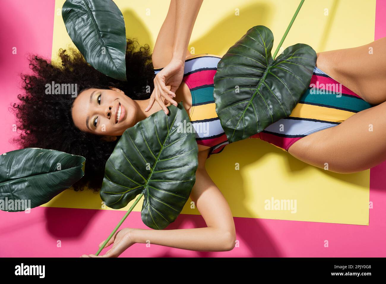 top view of cheerful african american woman in colorful swimsuit getting tan near green palm leaves on pink Stock Photo