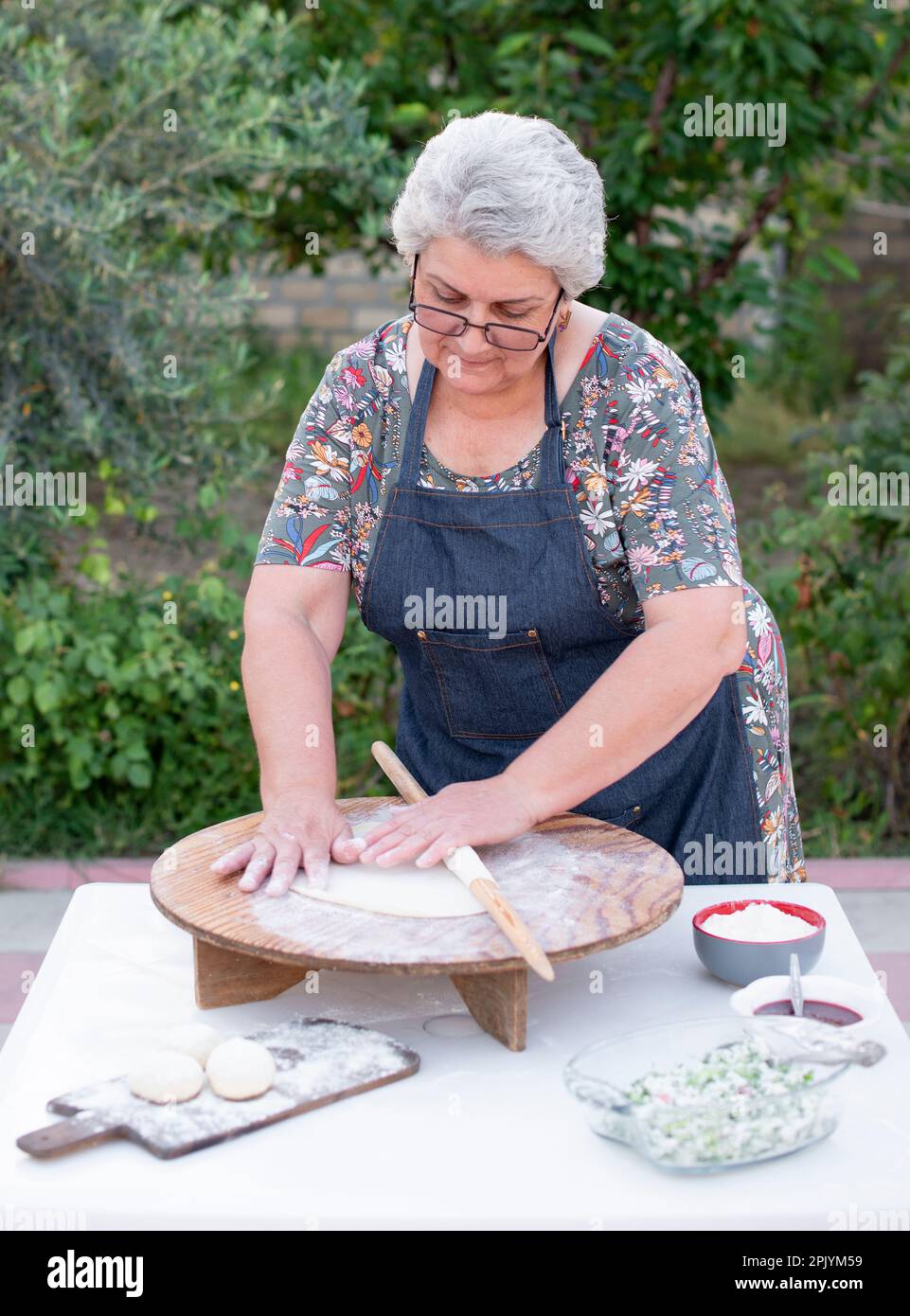 Old female cook, chef wearing apron looking dowm , touching dough, rolling, making pita. Stock Photo