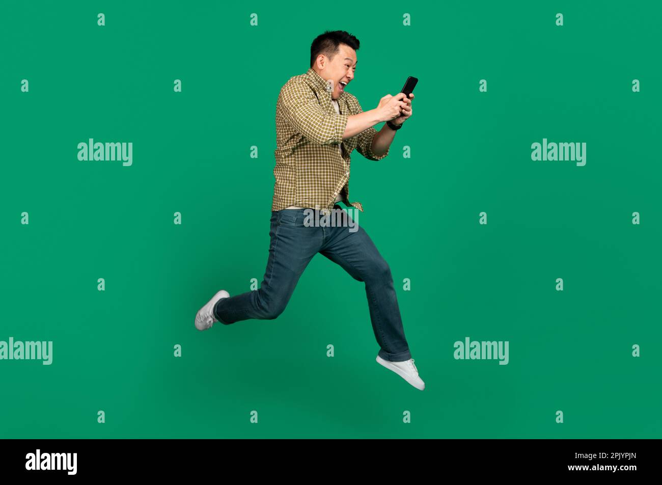 Full length of asian man with smartphone jumping up over green studio ...