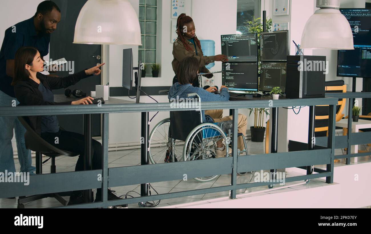 Asian coder with physical disability working with african american developer to create security firewall on terminasl window. IT engineer in wheelchair programming html script code. Stock Photo