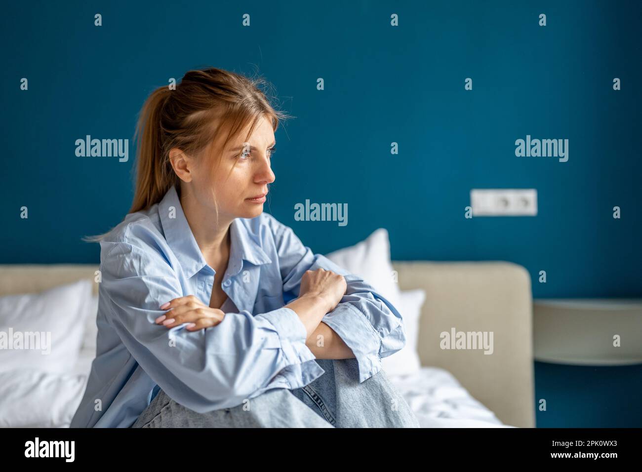 Woman exhausted by nervous stress looks devastated sits on bed with arms crossed feels hopelessness Stock Photo