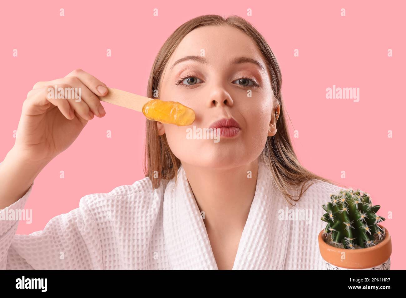 Young woman holding spatula with sugaring paste and cactus on pink background Stock Photo