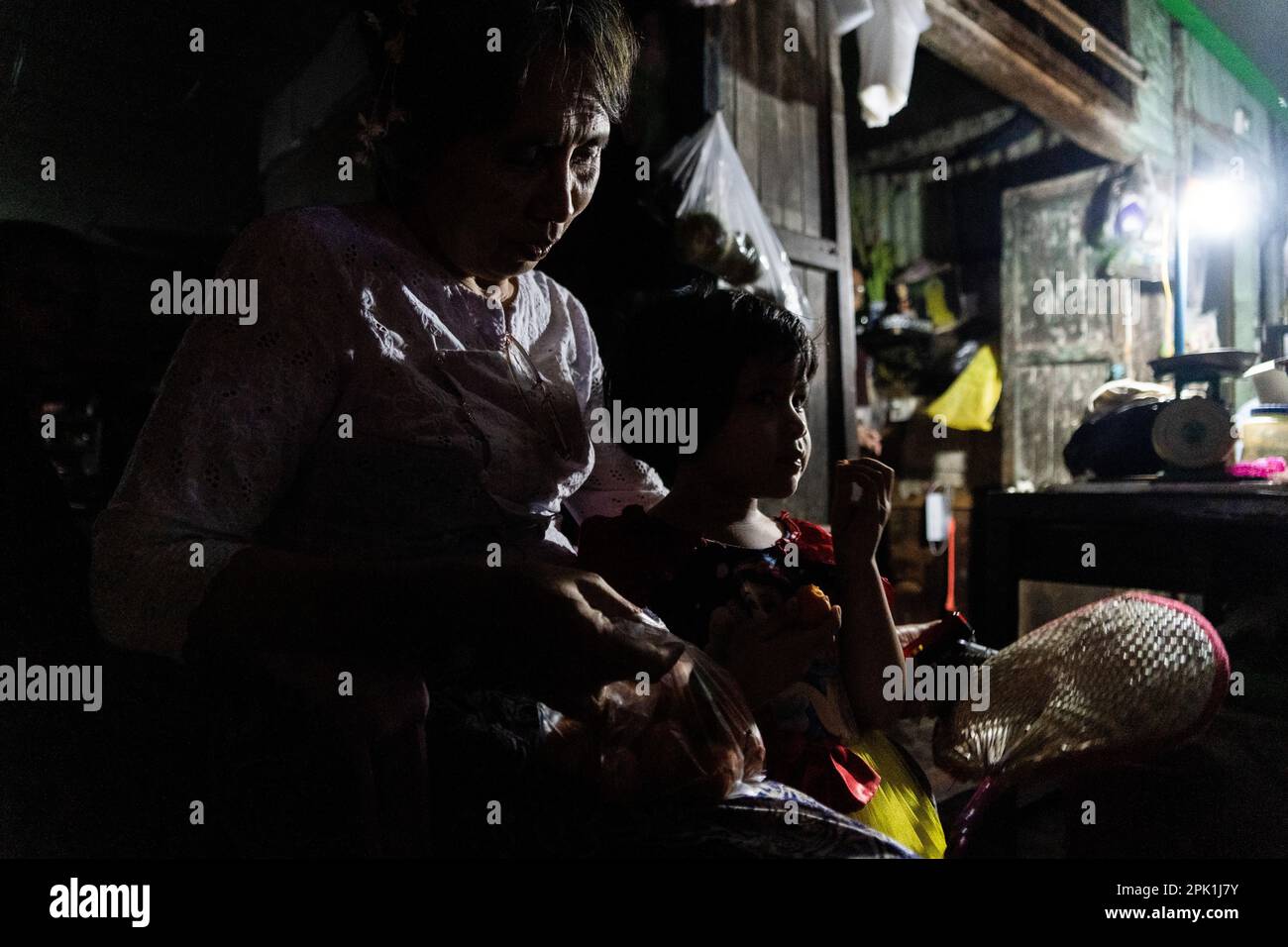 A young girl eats with her mother during a blackout at a shop on the street in Yangon. Due to soaring costs and the lack of hydropower during the dry season, the military junta (Tatmadaw) rations electricity throughout Yangon daily, leaving large sections of the city (townships) completely blacked out everyday. Daily life during the deadly civil war in Myanmar. On February 1, 2021, the military junta government (Tatmadaw) seized power by coup, jailing the democratically-elected government and plunging the country into an ongoing humanitarian crisis. Stock Photo