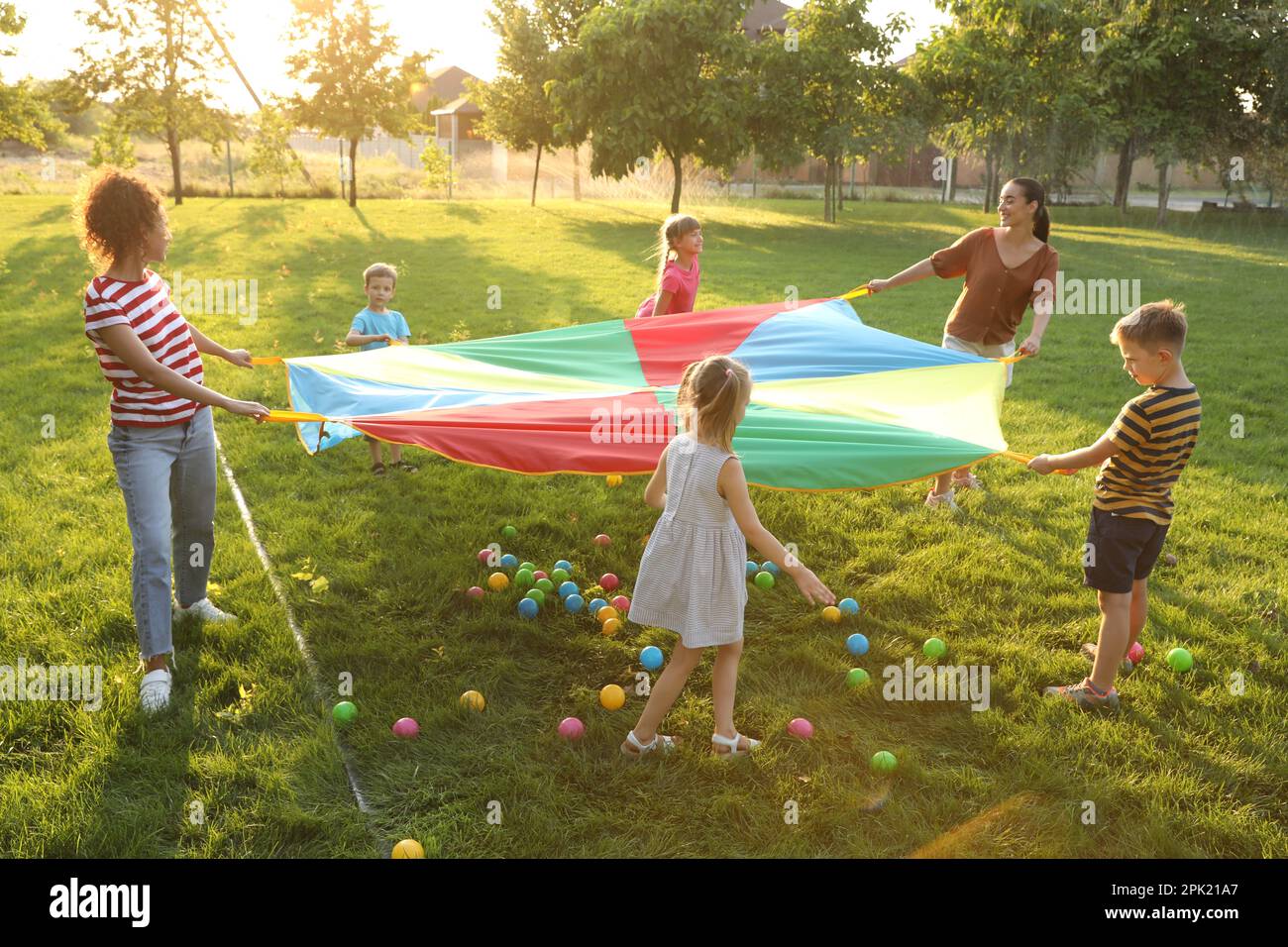 Group of children and teachers playing with rainbow playground parachute on green grass. Summer