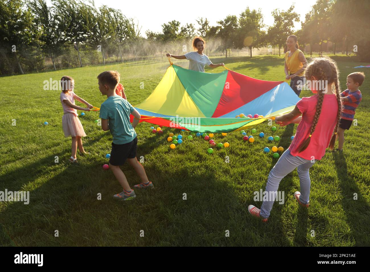 Group of children and teachers playing with rainbow playground parachute on green grass. Summer