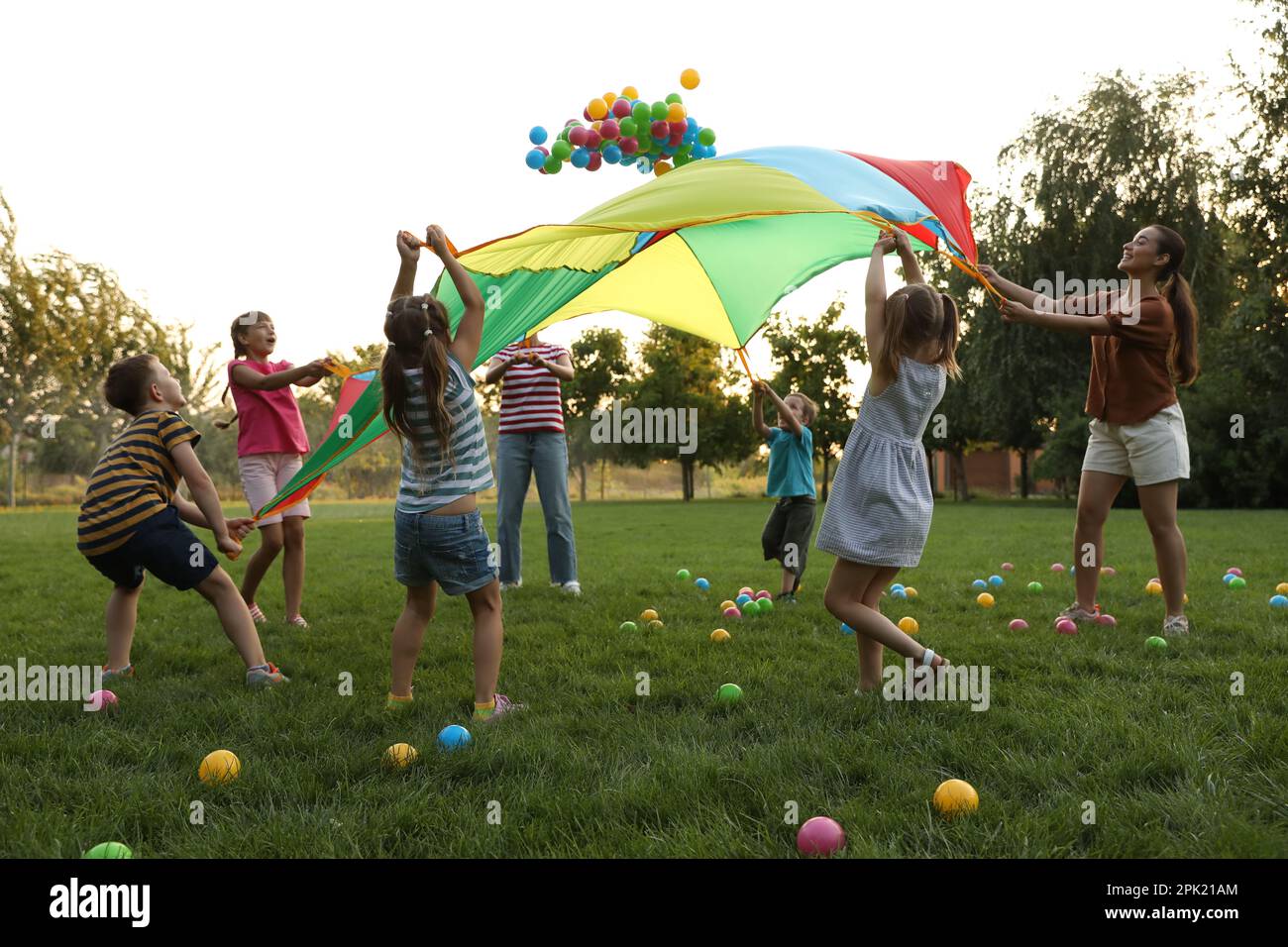 Group of children and teacher playing with rainbow playground parachute on green grass. Summer