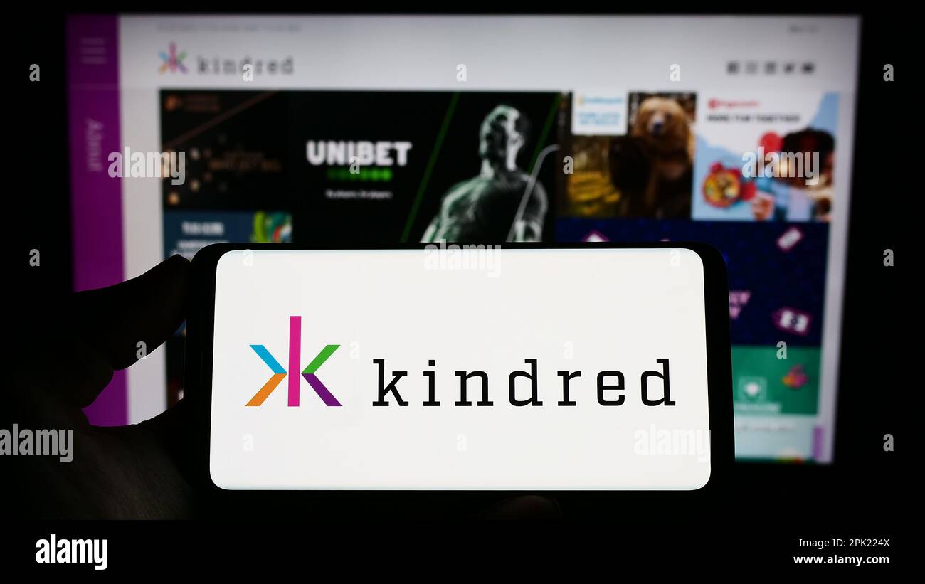 Person holding smartphone with logo of online gambling company Kindred Group plc on screen in front of website. Focus on phone display. Stock Photo
