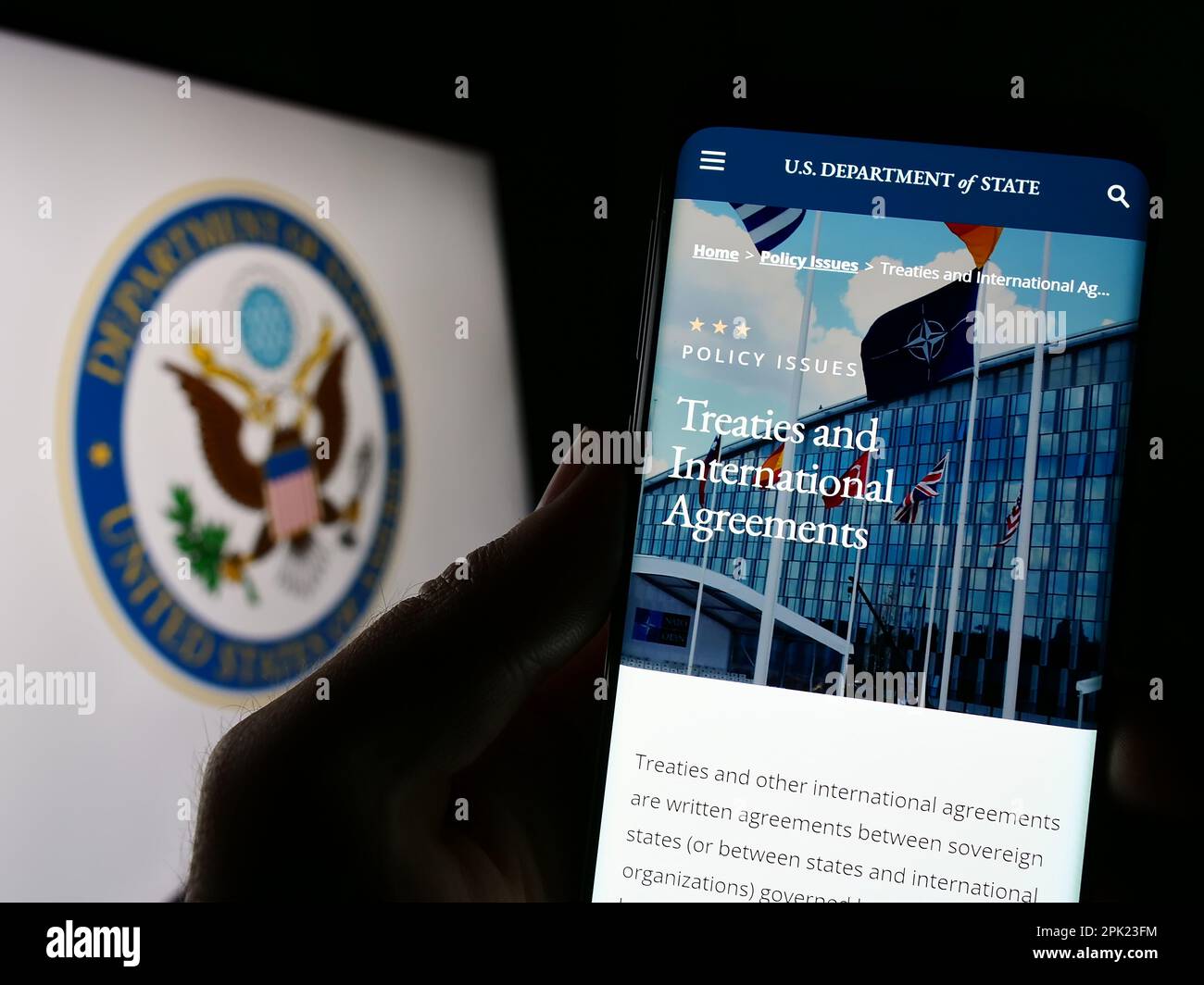 Person holding cellphone with web page of United States Department of State (DOS) on screen in front of logo. Focus on center of phone display. Stock Photo