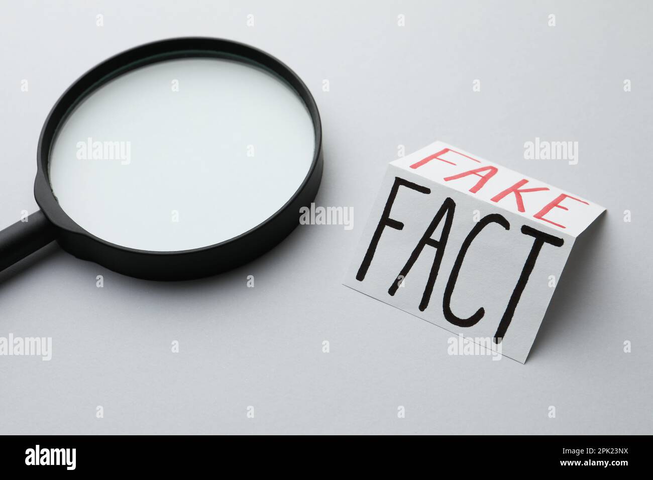 Magnifying glass near sheet of paper with words Fact and Fake on light