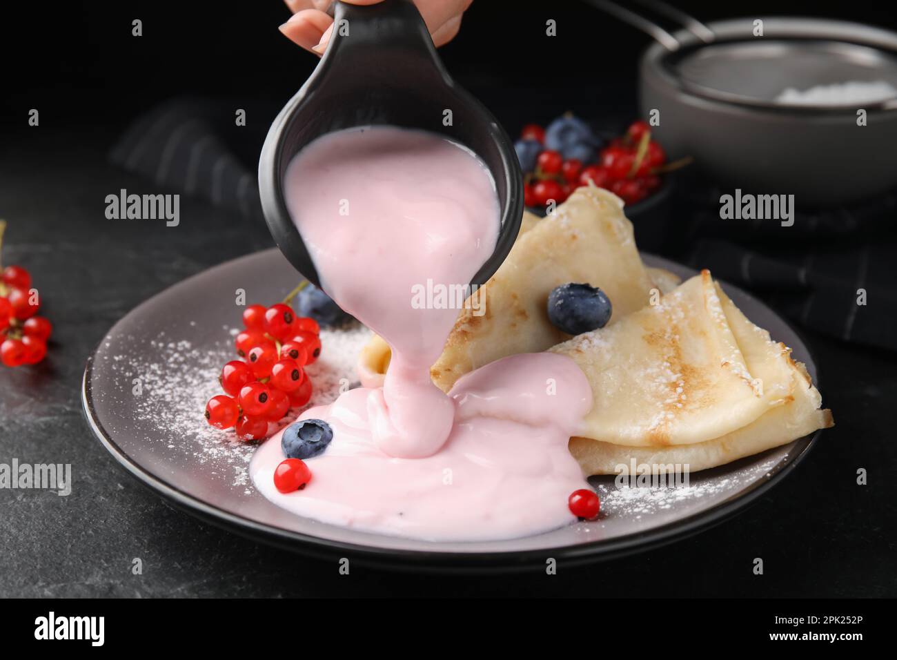 Woman pouring natural yogurt onto crepes with blueberries and red currants at table, closeup Stock Photo