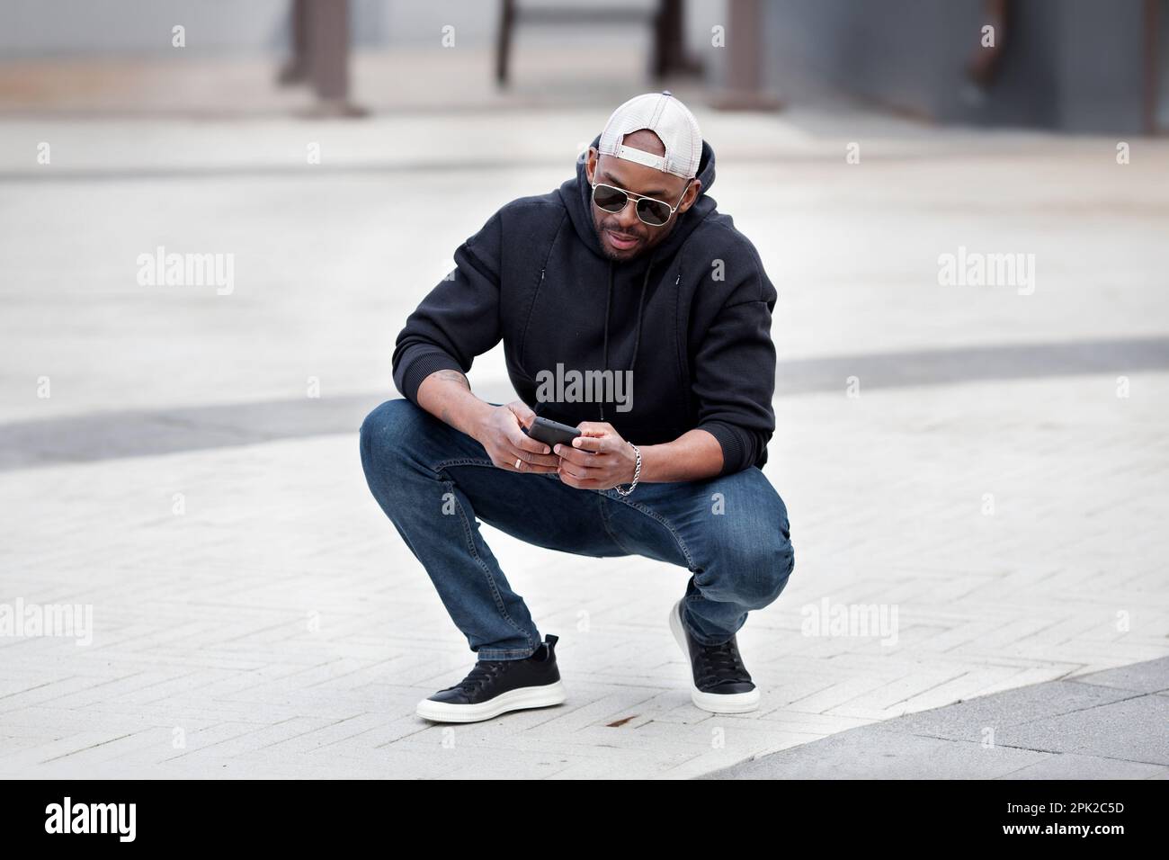 Cool African American man using smartphone squatting on city street wears black hoodie jeans and sneakers baseball cap and sunglasses. Stylish guy Stock Photo Alamy