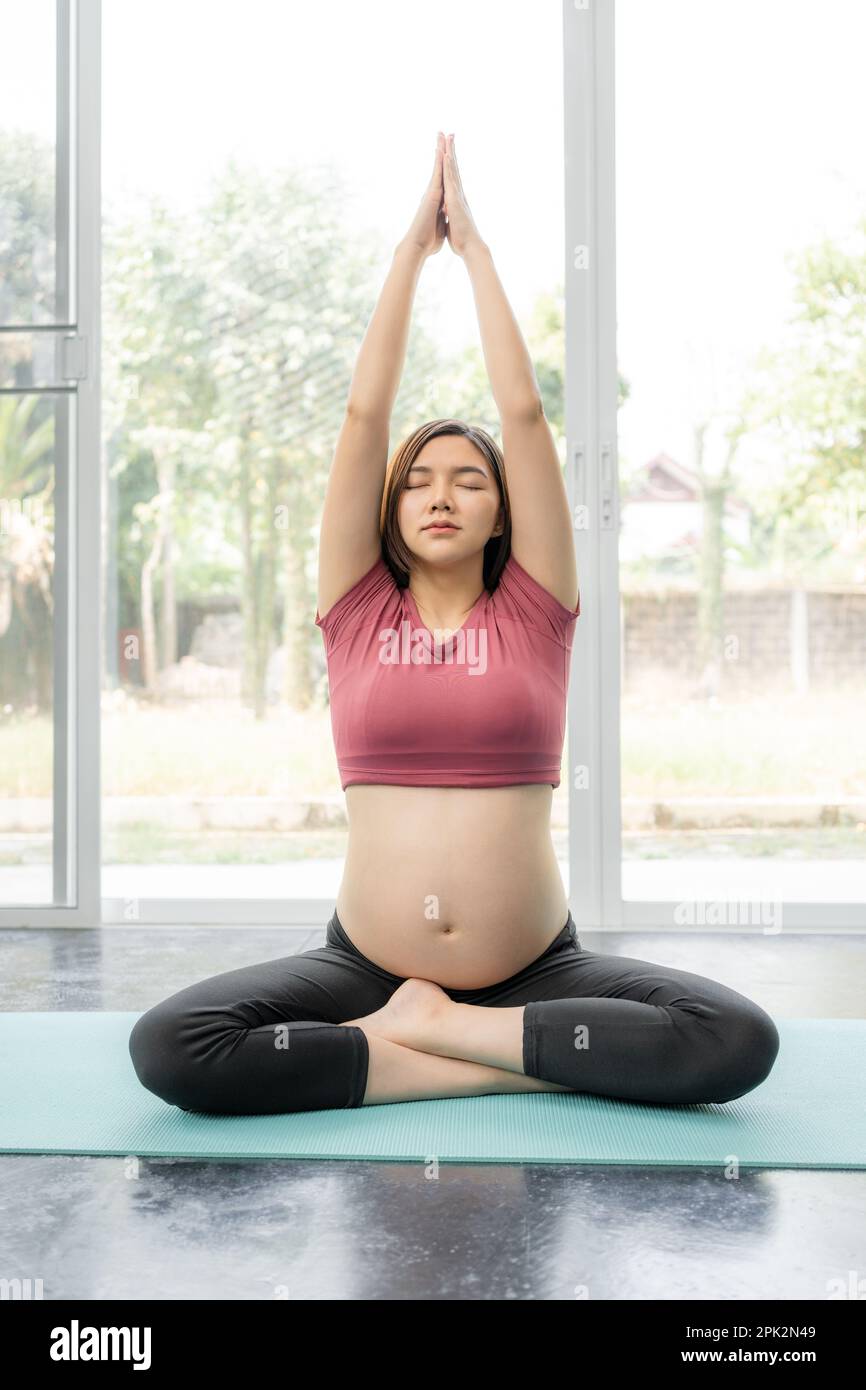 Happy young pregnant Asian woman in fitness clothes sitting cross legs and stretching her arms upward during her yoga exercise practice in her home li Stock Photo