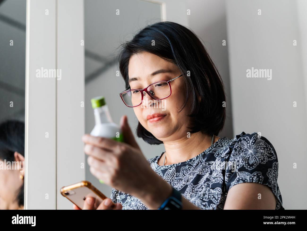 Asian woman is reading medicine information from her smartphone, her hand is holding a liquid medicine bottle, eyes looking at her cellphone. Asian mi Stock Photo