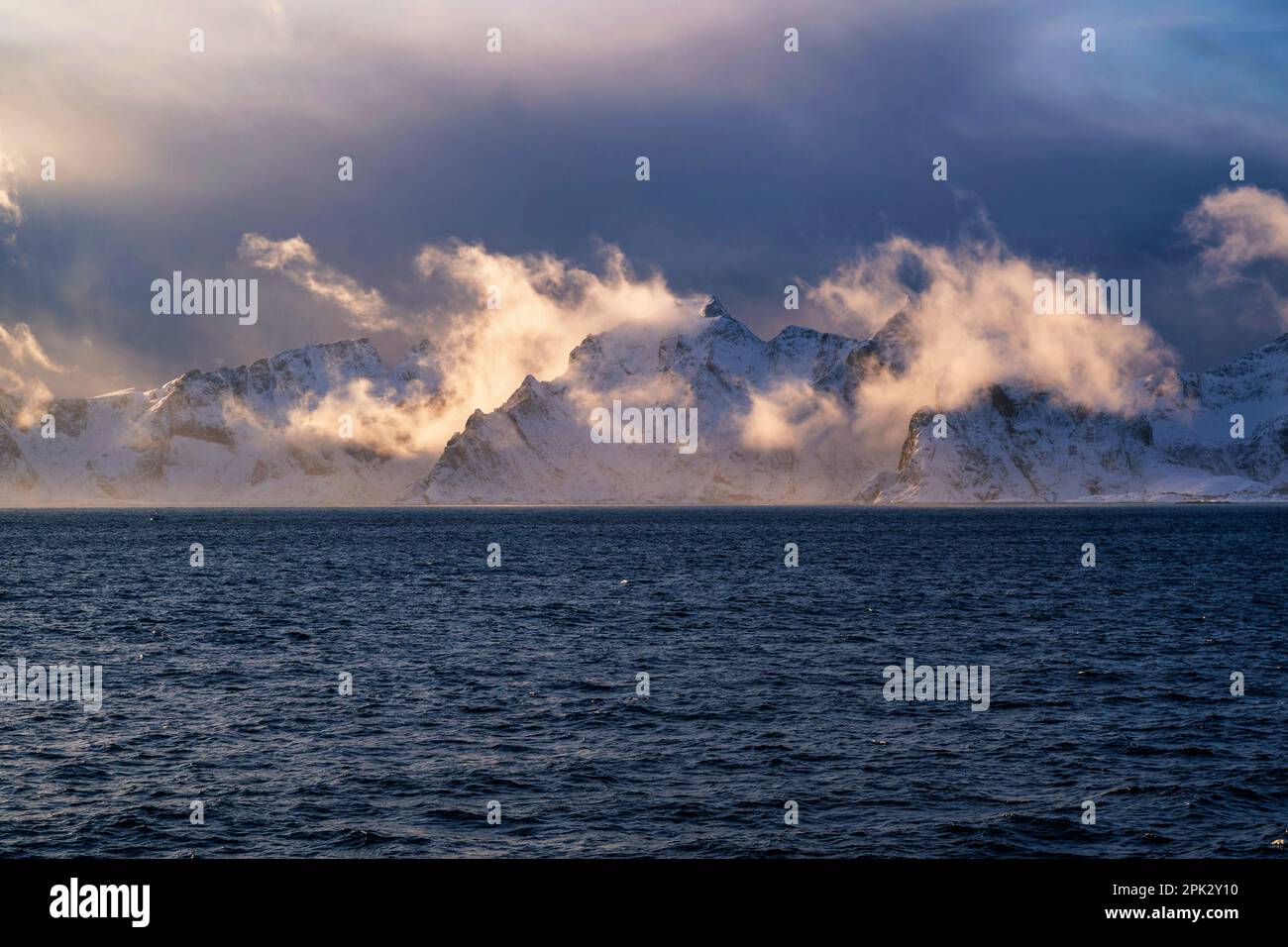 Gorgeous view to snowy rocky mountain on dramatic background of cloudy sky illumination by setting sun from boat. Lofoten, Norway. Stock Photo