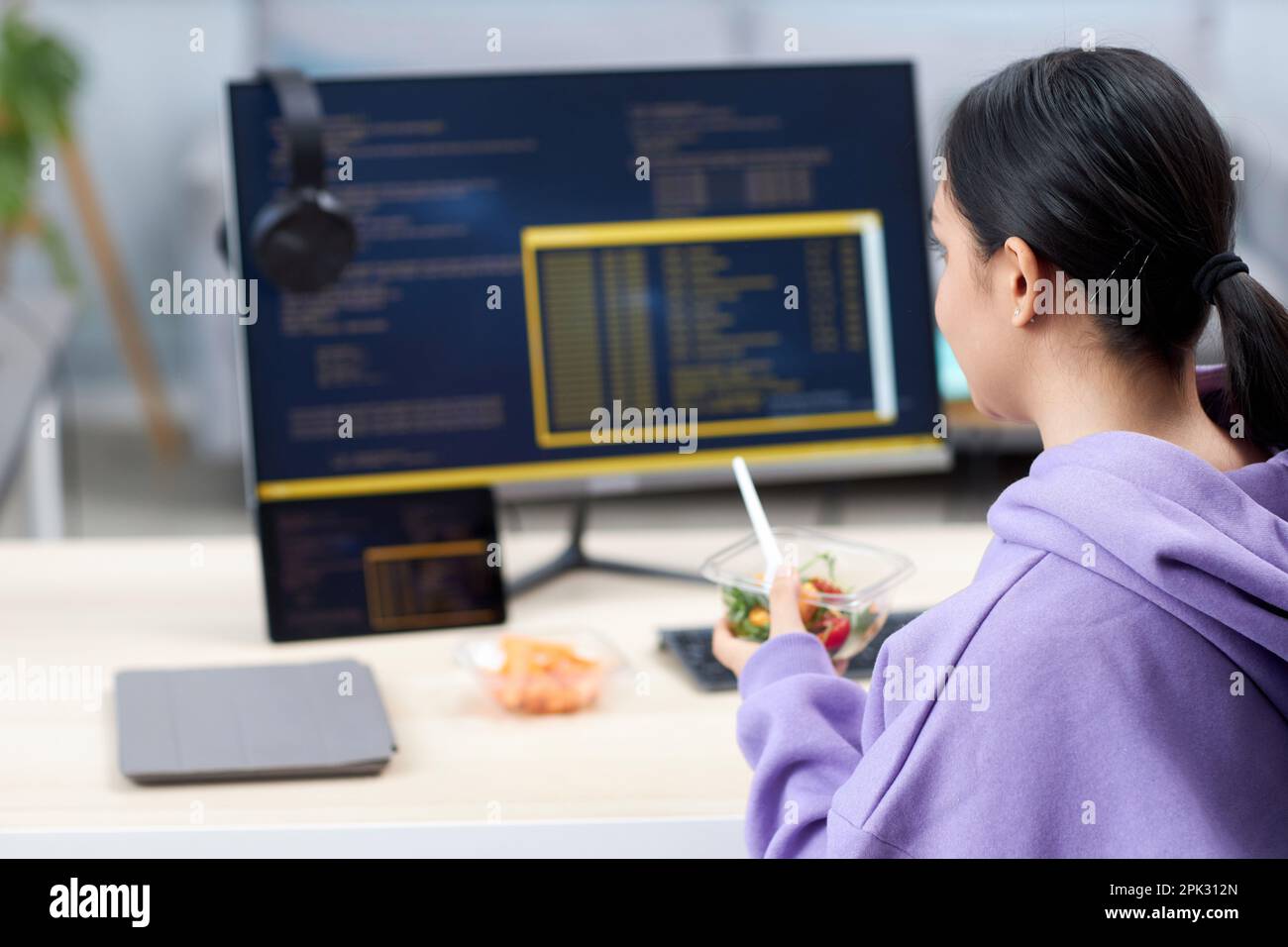 Back view of female computer programmer eating takeout lunch at workplace while writing code, copy space Stock Photo