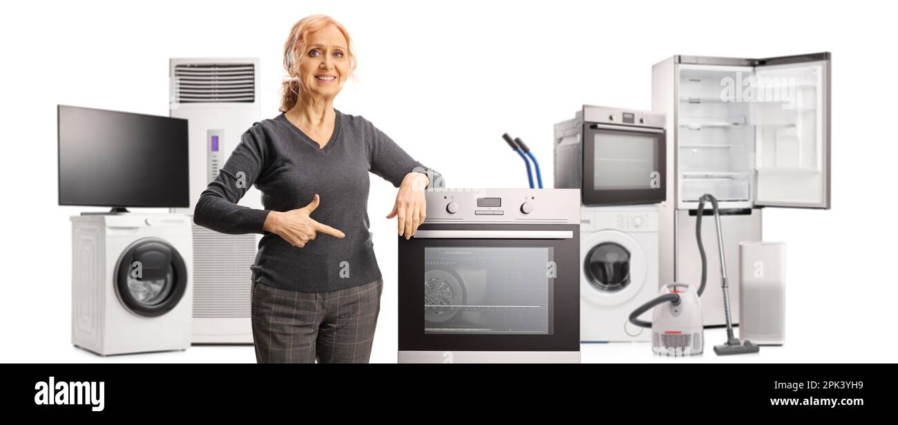Smiling mature woman pointing at an oven and posing with other home applances in the back isolated on white background Stock Photo
