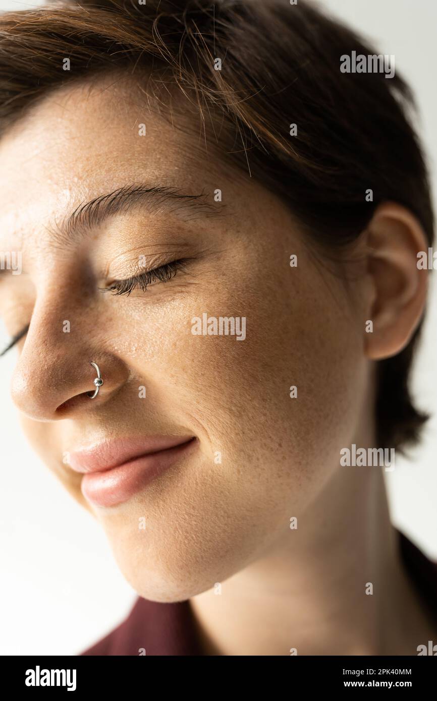 close up portrait of carefree woman with freckles and nose piercing smiling with closed eyes on grey background,stock image Stock Photo