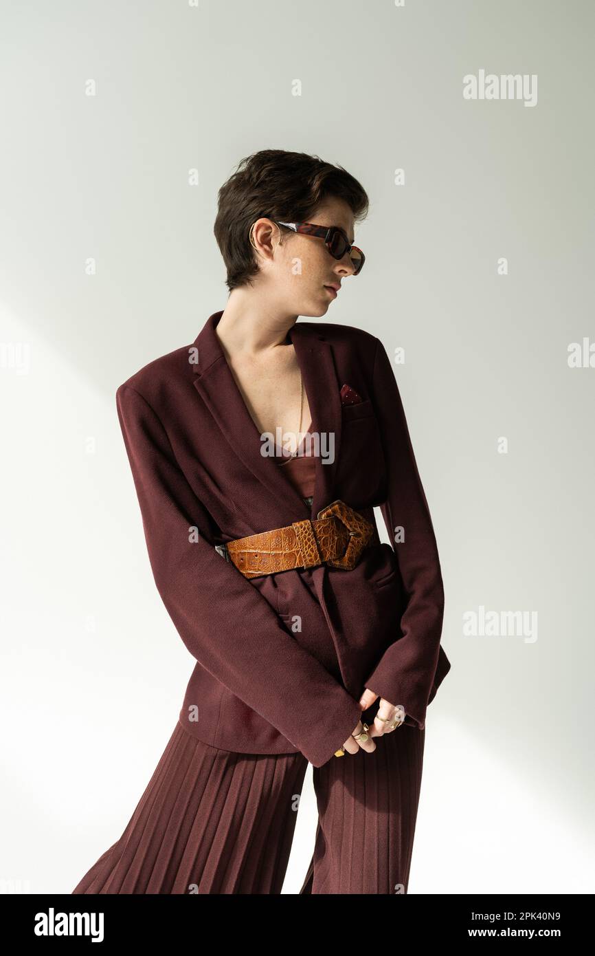 brunette woman in sunglasses and pantsuit with leather belt on grey background,stock image Stock Photo