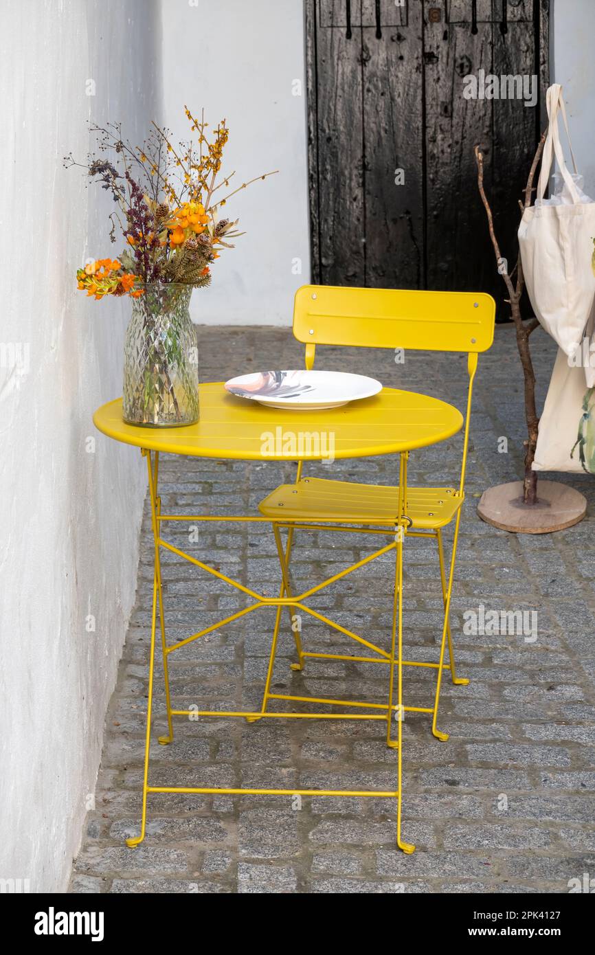 Cute corner with yellow iron table and chair on which there is a plate and a vase with flowers. Stock Photo