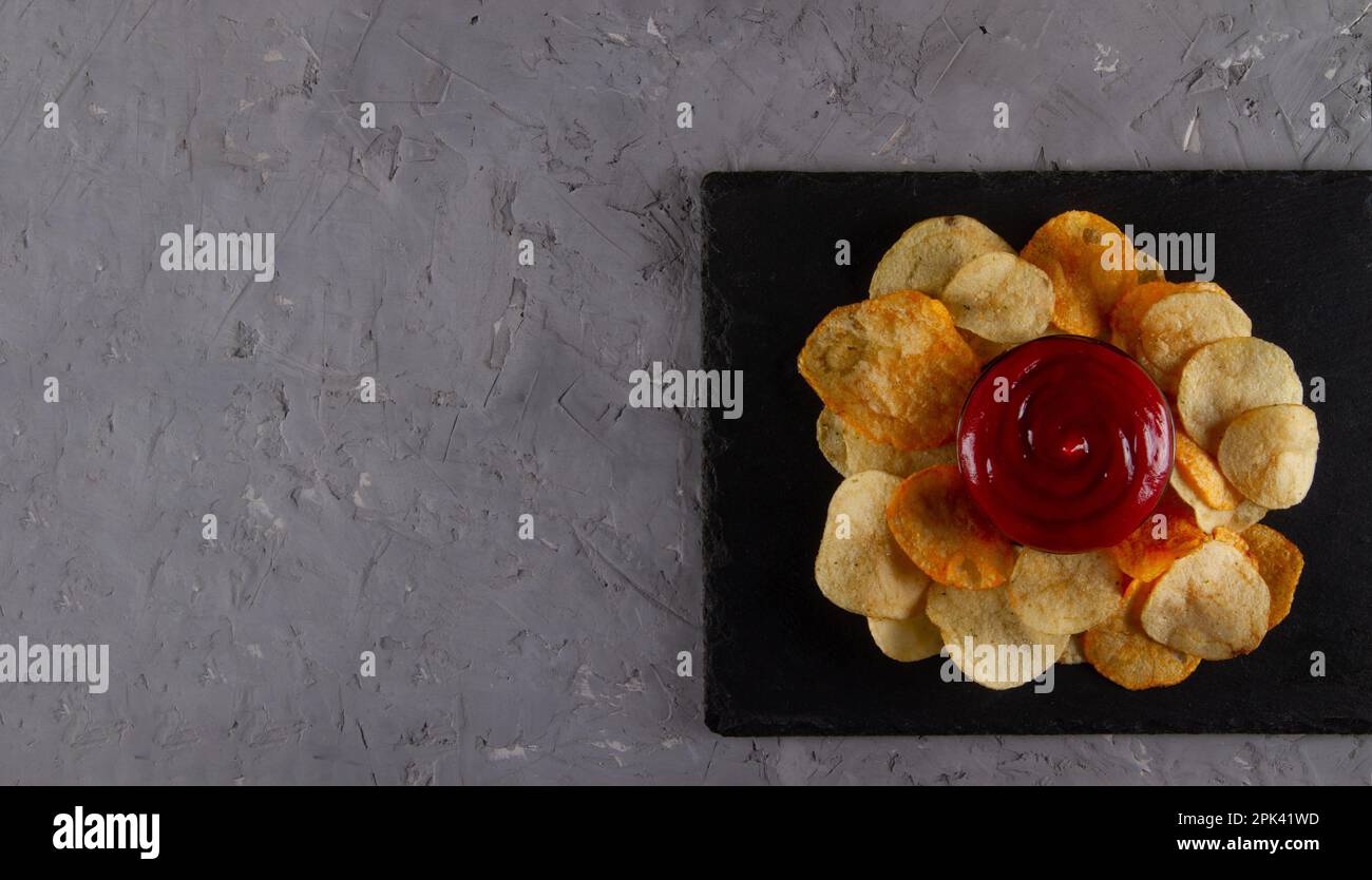 Top view of heap of potato chips with red dipping sauce on black slate serving board on grey plaster table. Gourmet Potato Chips Display Stock Photo