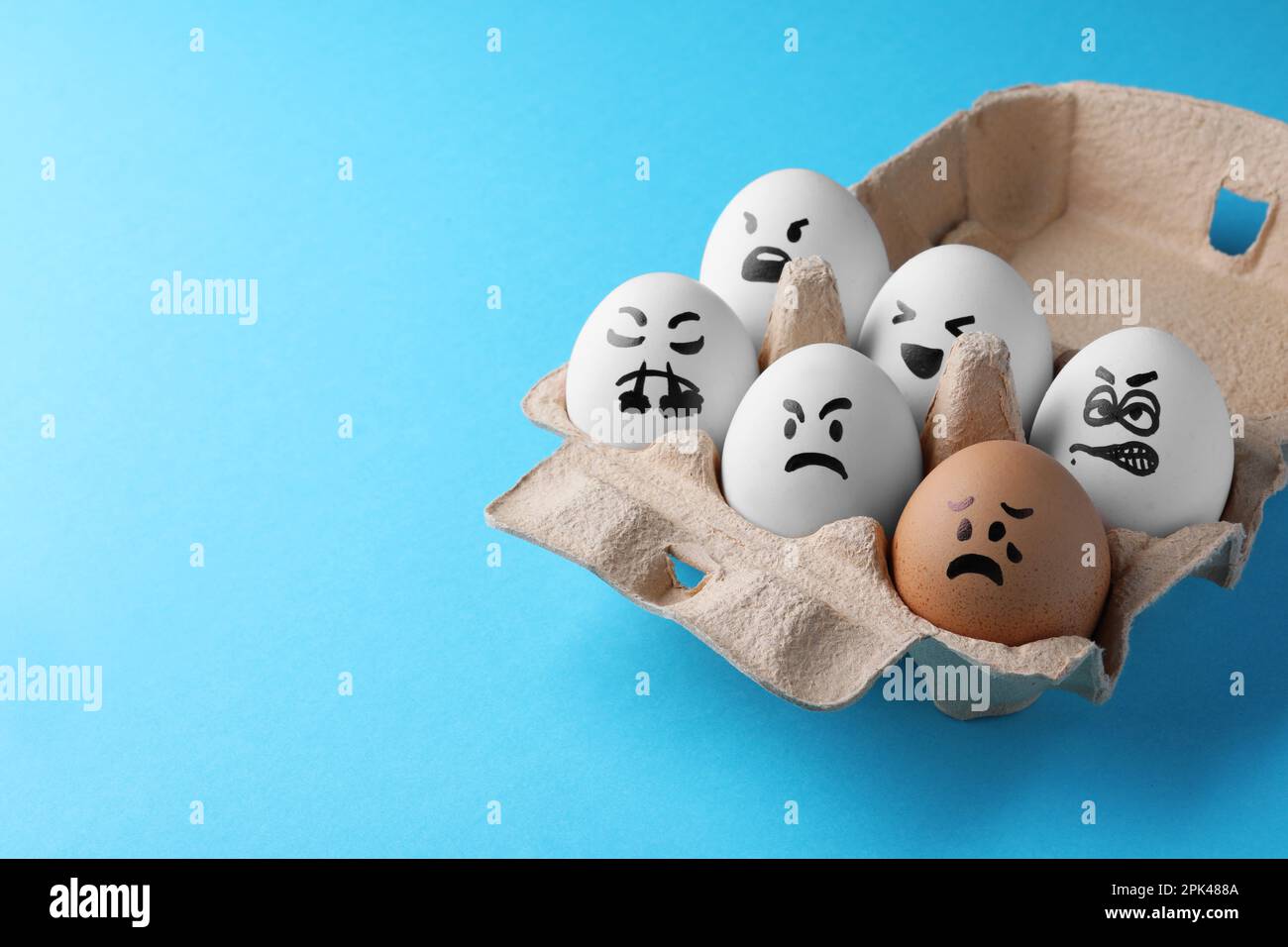 Brown egg with upset face among aggressively disposed white ones in carton box on turquoise background, space for text. Bullying concept Stock Photo