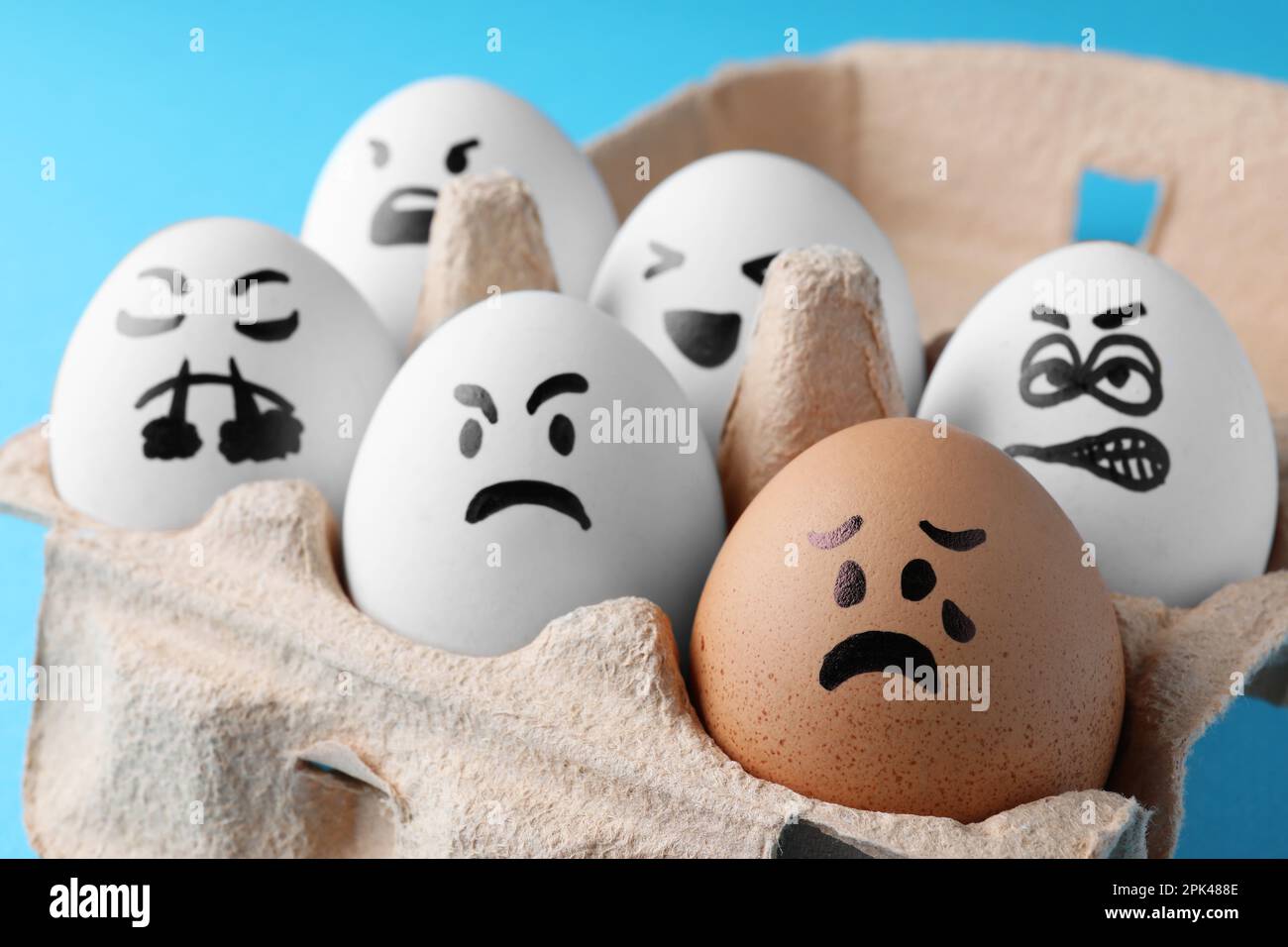 Brown egg with upset face among aggressively disposed white ones in carton box on turquoise background, closeup. Bullying concept Stock Photo