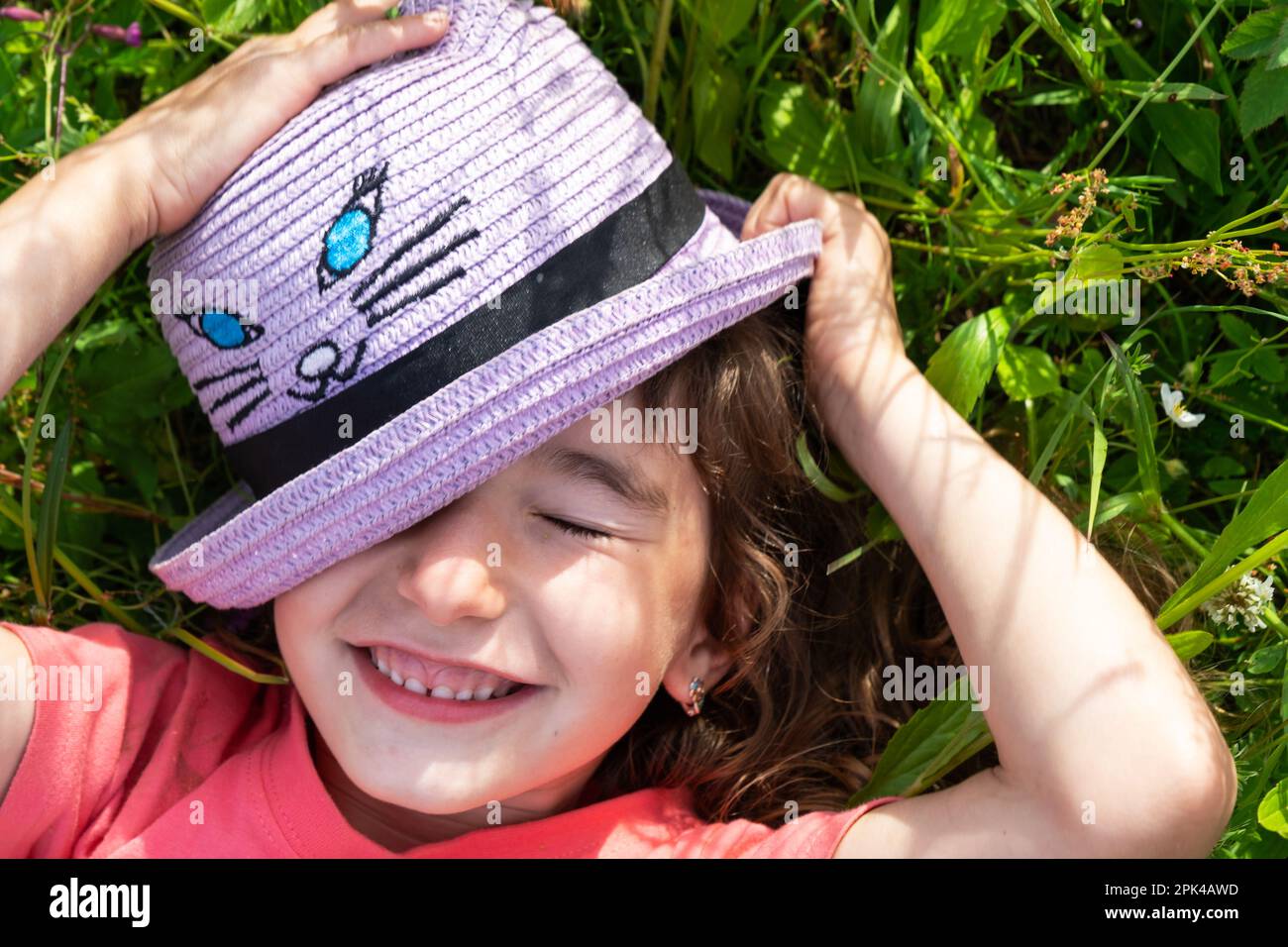 Portrait of a child in a hat with his face covered in summer lying in the grass and wildflowers. Hat with eyes and ears like a cat, summer time, freed Stock Photo