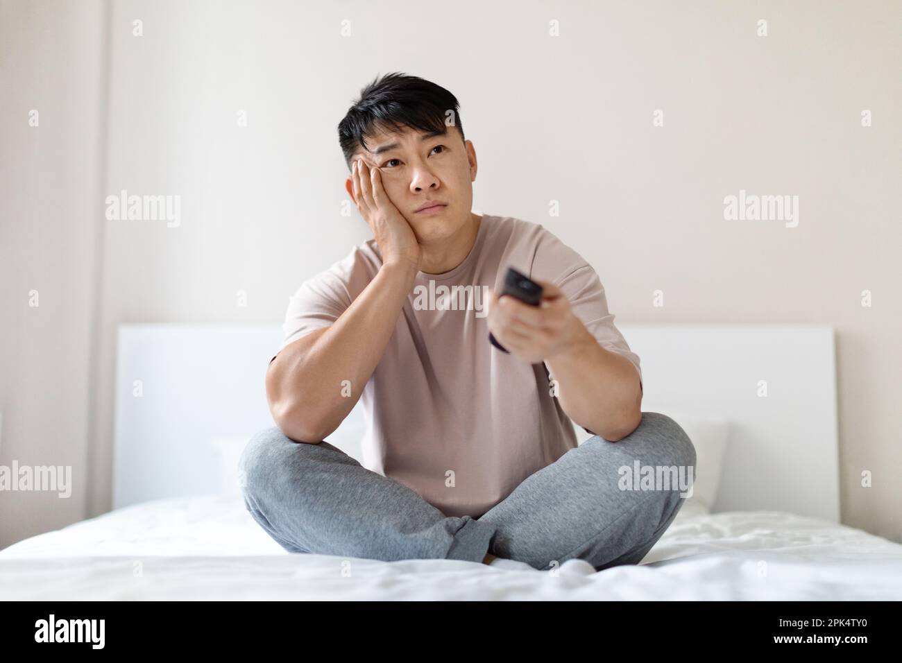 Dull asian man sitting on bed at home, holding remote Stock Photo