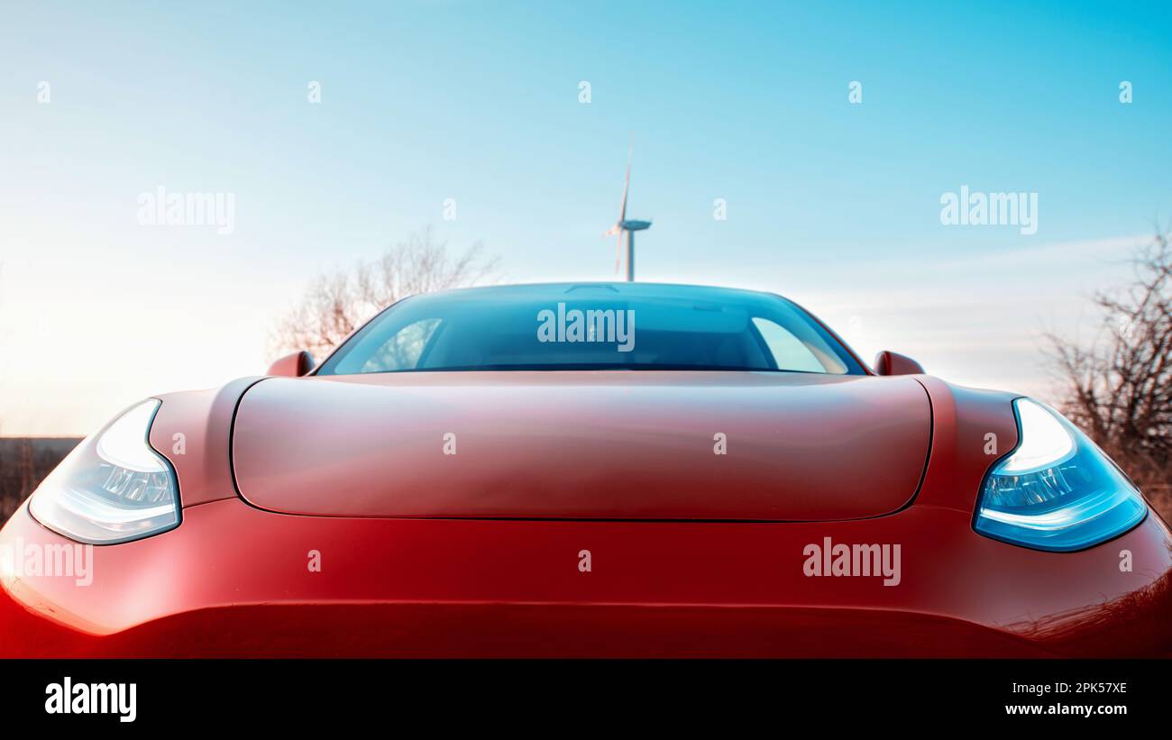 Close front view of a parked red electric car. Wind turbine on the