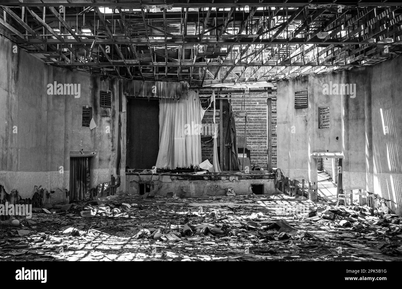 Black and White Monochrome Abandoned and Decaying Building with a Movie Theater Screen with trash on the floor and sunlight beams through exposed sky Stock Photo
