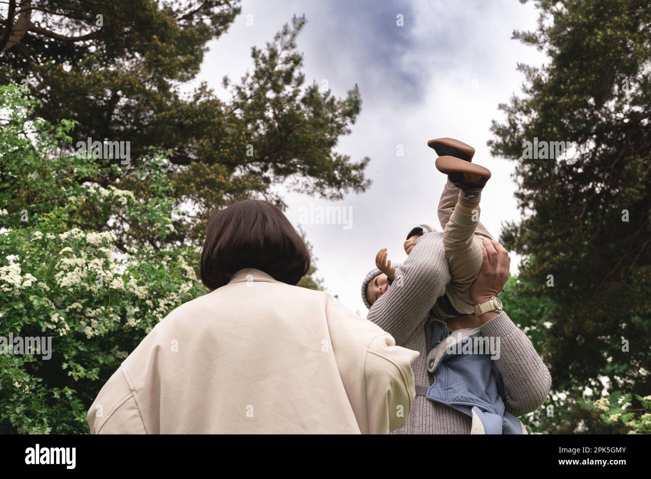 Low perspective photo of multicultural family playing with baby, lifting, throwing infant up in air in nature. Unrecognizable father is holding kid in Stock Photo