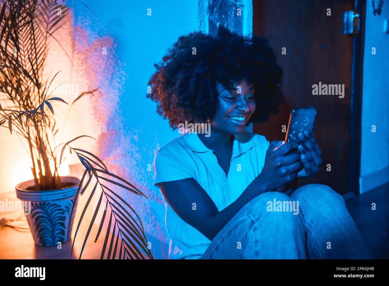Woman with afro hair sitting at home on the floor looking at the phone at night with a blue light Stock Photo