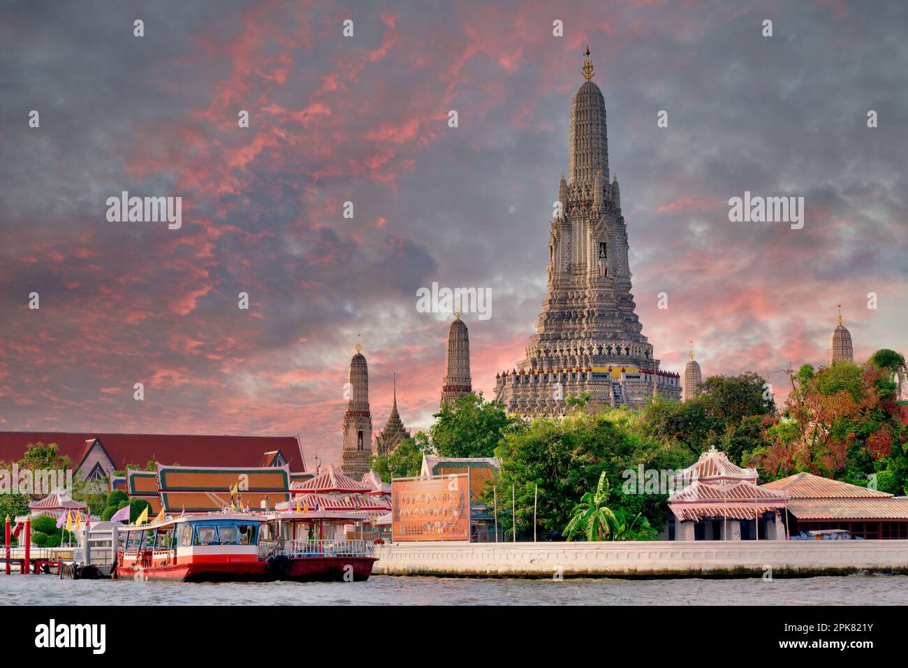 Wat Arun, a unique, landmark Buddhist temple in Bangkok, Thailand, under a colorful sky; in front: the Chao Phraya River and boats anchored at a pier Stock Photo
