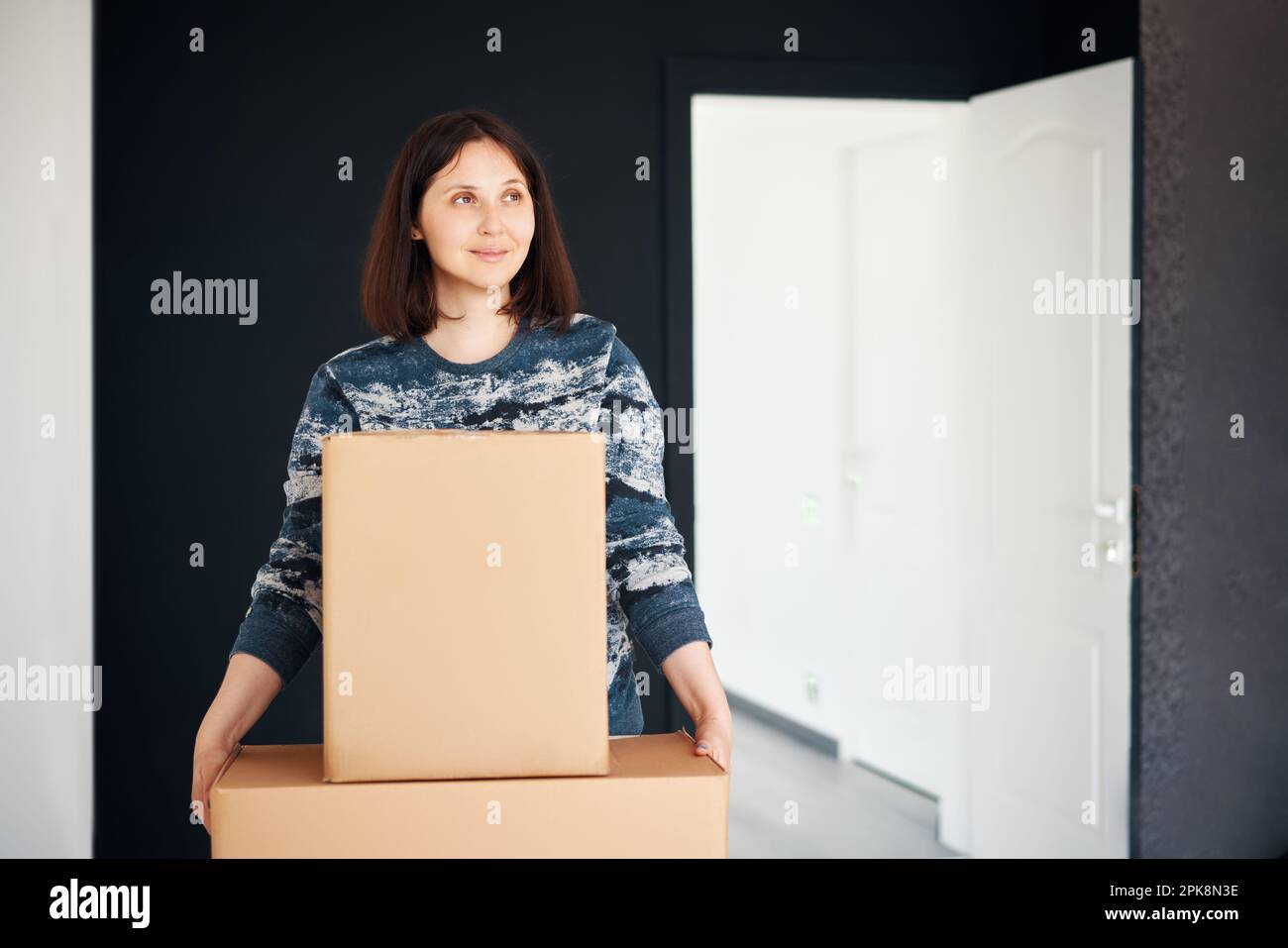 Young smiling woman moving into new apartment carrying cardboard boxes. Relocation concept Stock Photo