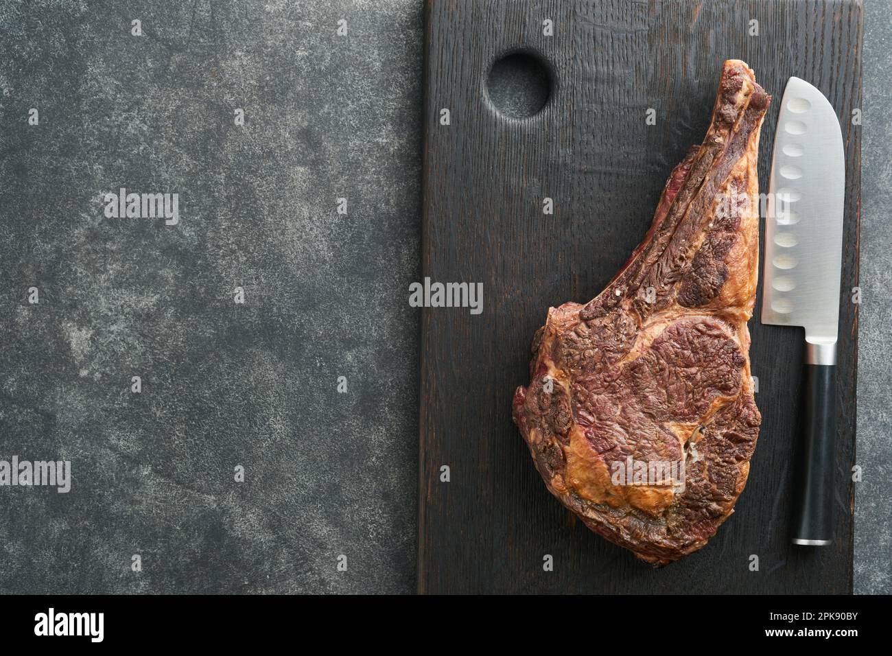 Tomahawk steak. Sliced grilled tomahawk beef steak with baked cherry tomatoes, herbs and salt on old wooden background. Preparing to grill.  Top view Stock Photo
