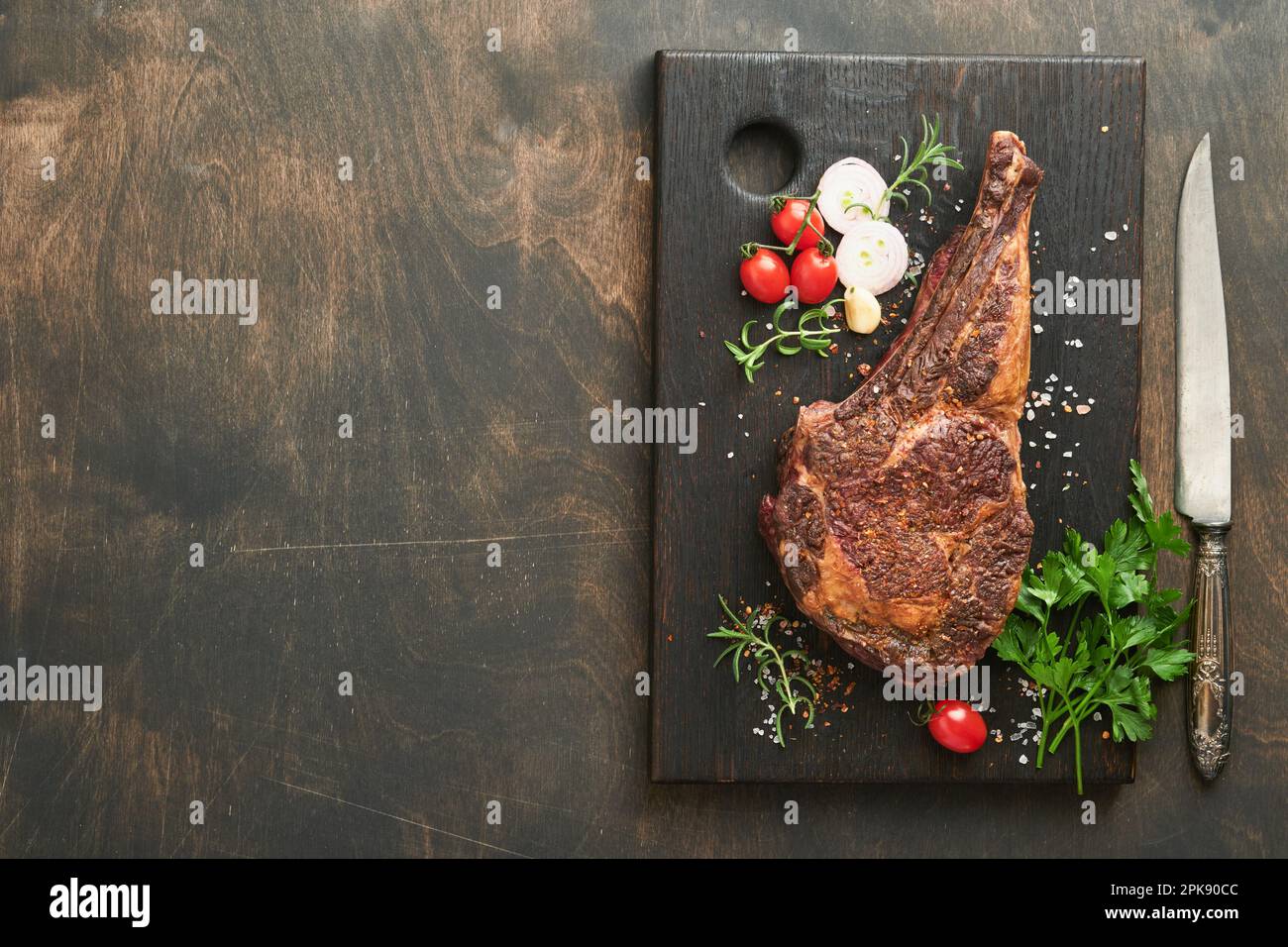 Tomahawk steak. Sliced grilled tomahawk beef steak with baked cherry tomatoes, herbs and salt on old wooden background. Preparing to grill.  Top view Stock Photo