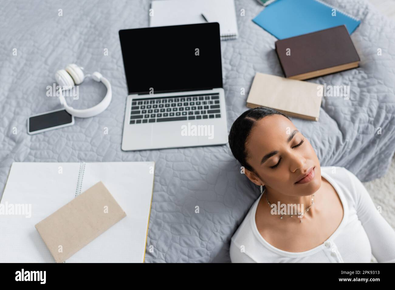 top view of african american student with closed eyes resting near laptop and books on bed,stock image Stock Photo