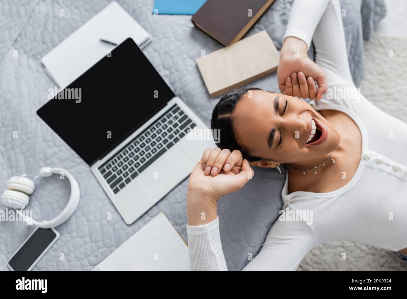 top view of tired african american student with closed eyes yawning near laptop and books on bed,stock image Stock Photo
