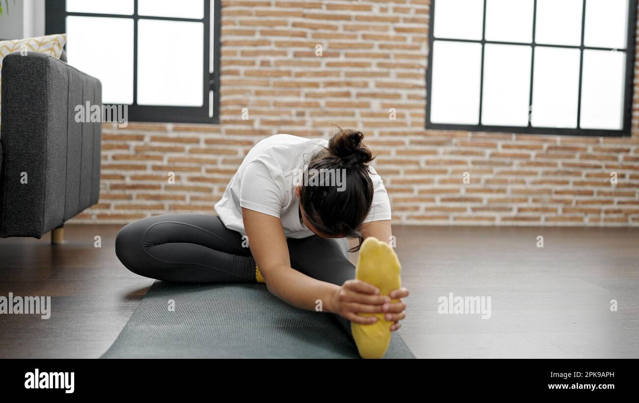Young beautiful hispanic woman sitting on floor stretching legs at home ...