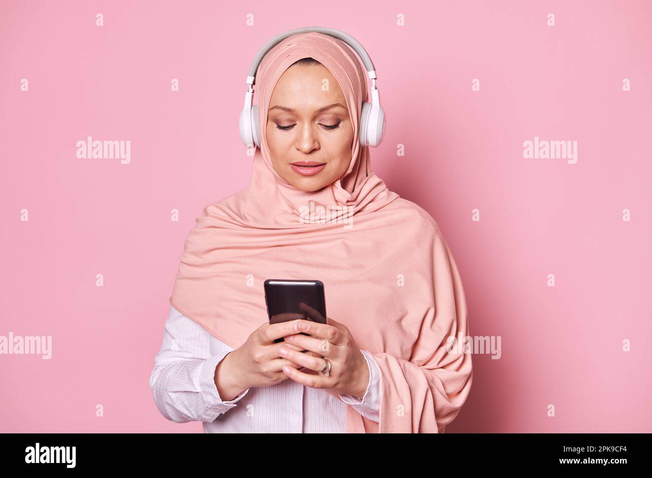 Close-up portrait of a Middle-Eastern Muslim woman in wireless headphones, using mobile phone against pink background Stock Photo