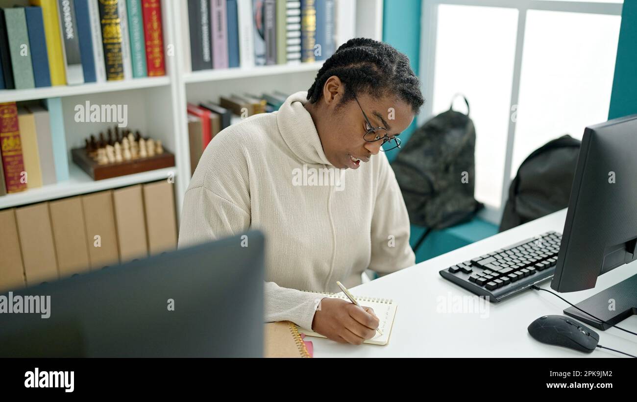 African american woman student using computer writing notes at library ...