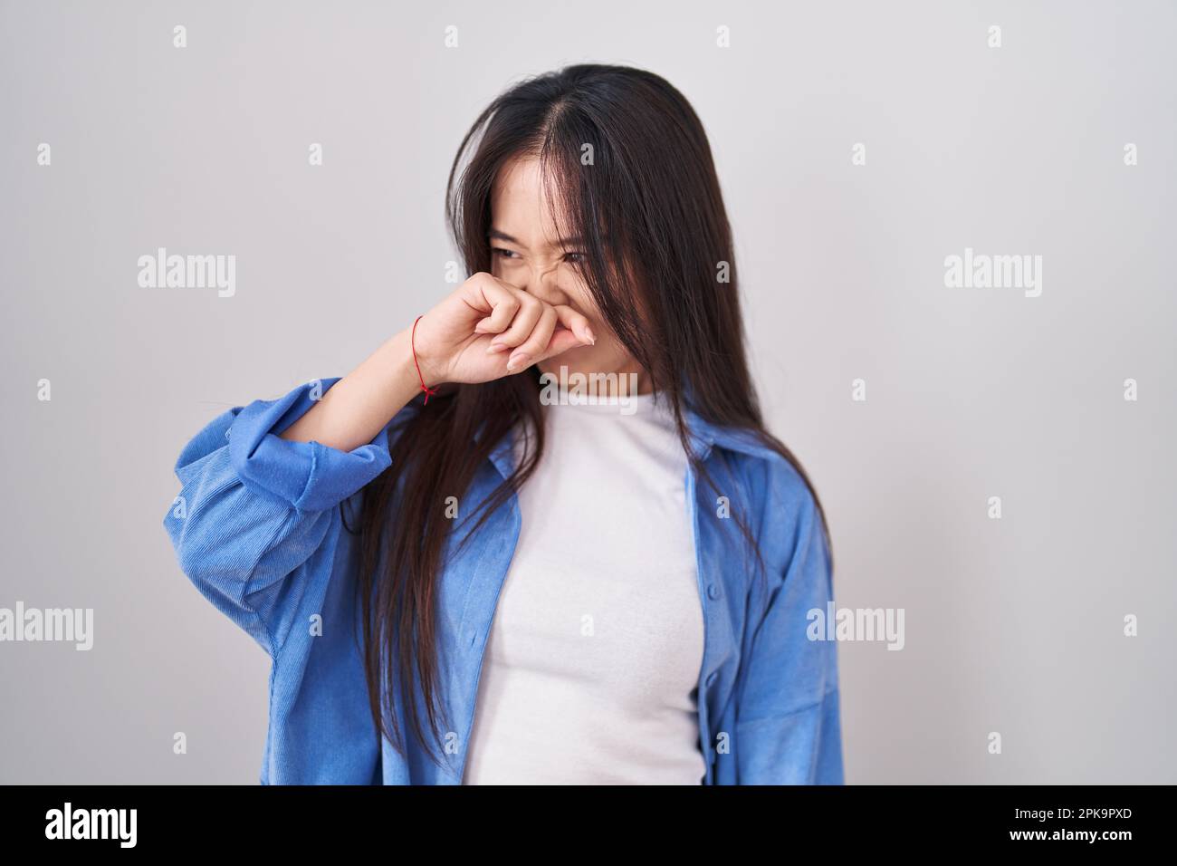 Young chinese woman standing over white background smelling something stinky and disgusting, intolerable smell, holding breath with fingers on nose. b Stock Photo