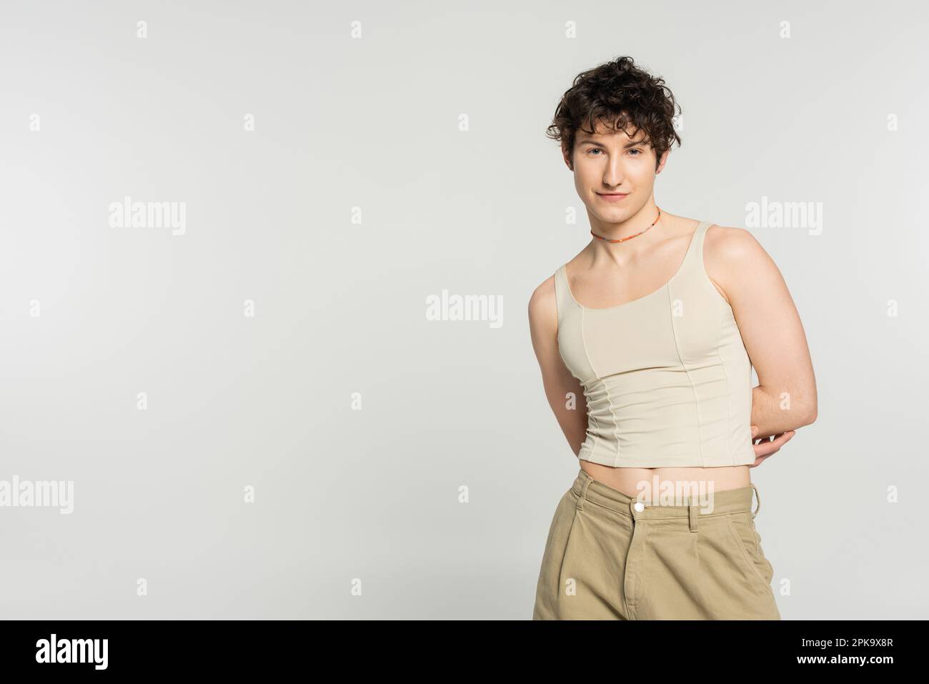 young bigender model in tank top and pants standing with hands behind ...