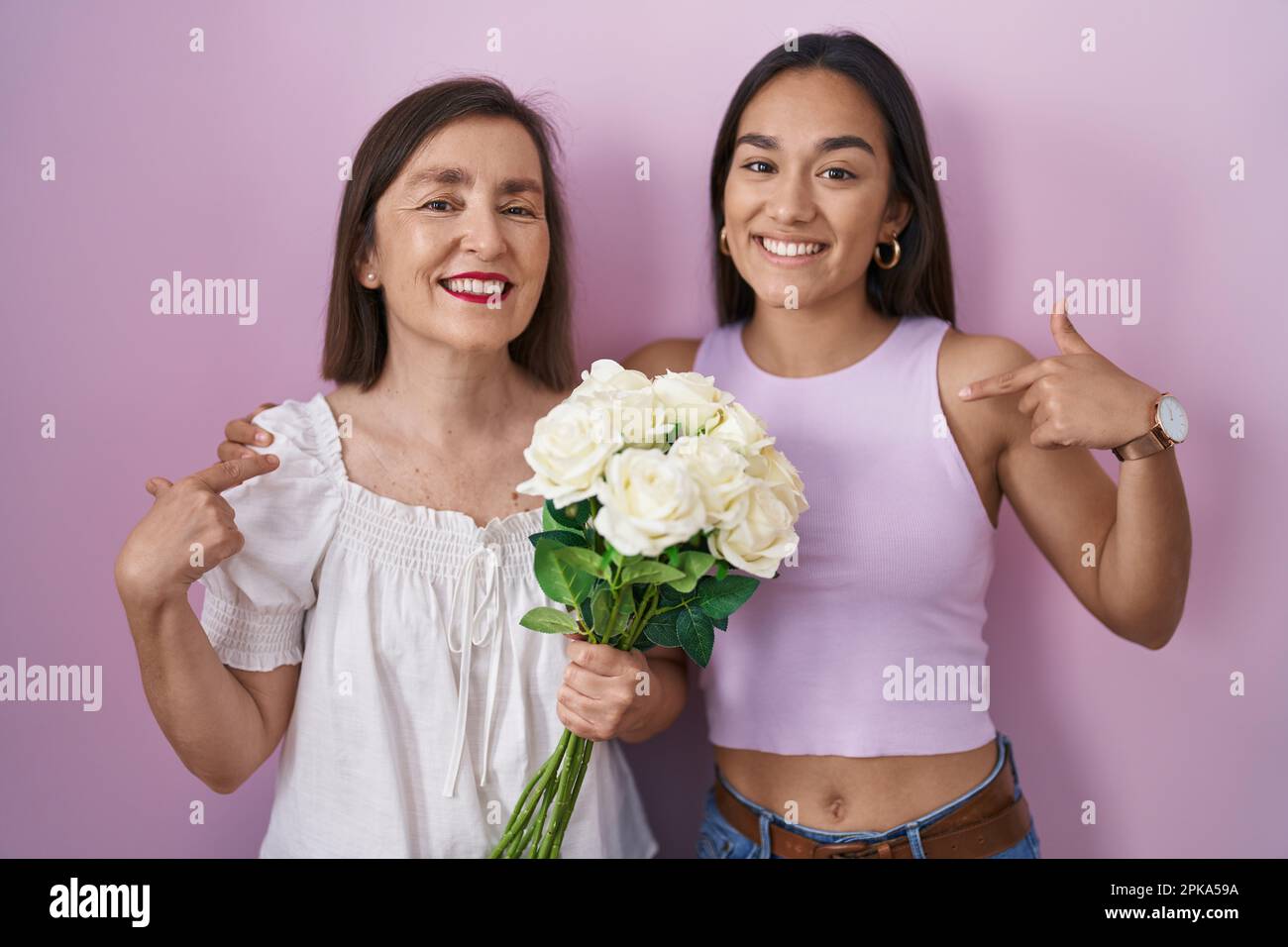 Hispanic mother and daughter holding bouquet of white flowers looking confident with smile on face, pointing oneself with fingers proud and happy. Stock Photo