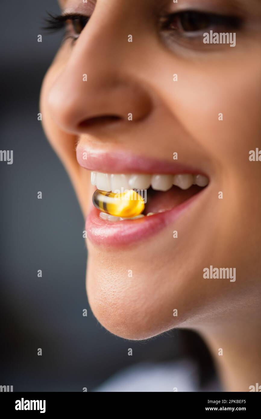 Multiracial Happy Young woman holding yellow pill mouth Between Teeth in natural fish oil Omega 3 vitamins pill standing by window at home Close-Up Stock Photo