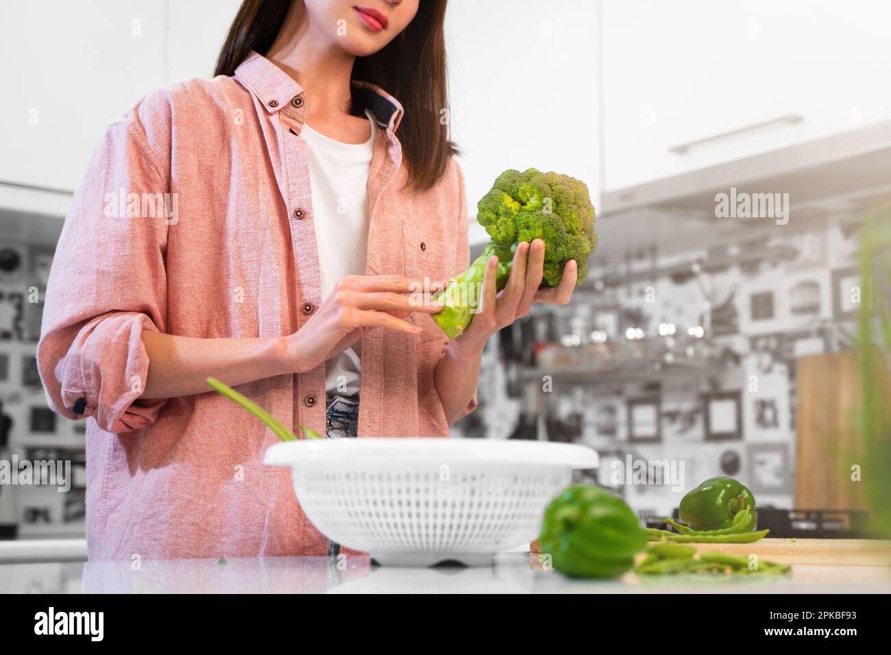 A Young woman in oversize pink shirt stands in the home kitchen, holding green broccoli in hands, going to cook a vegetarian diet food. The concept Stock Photo