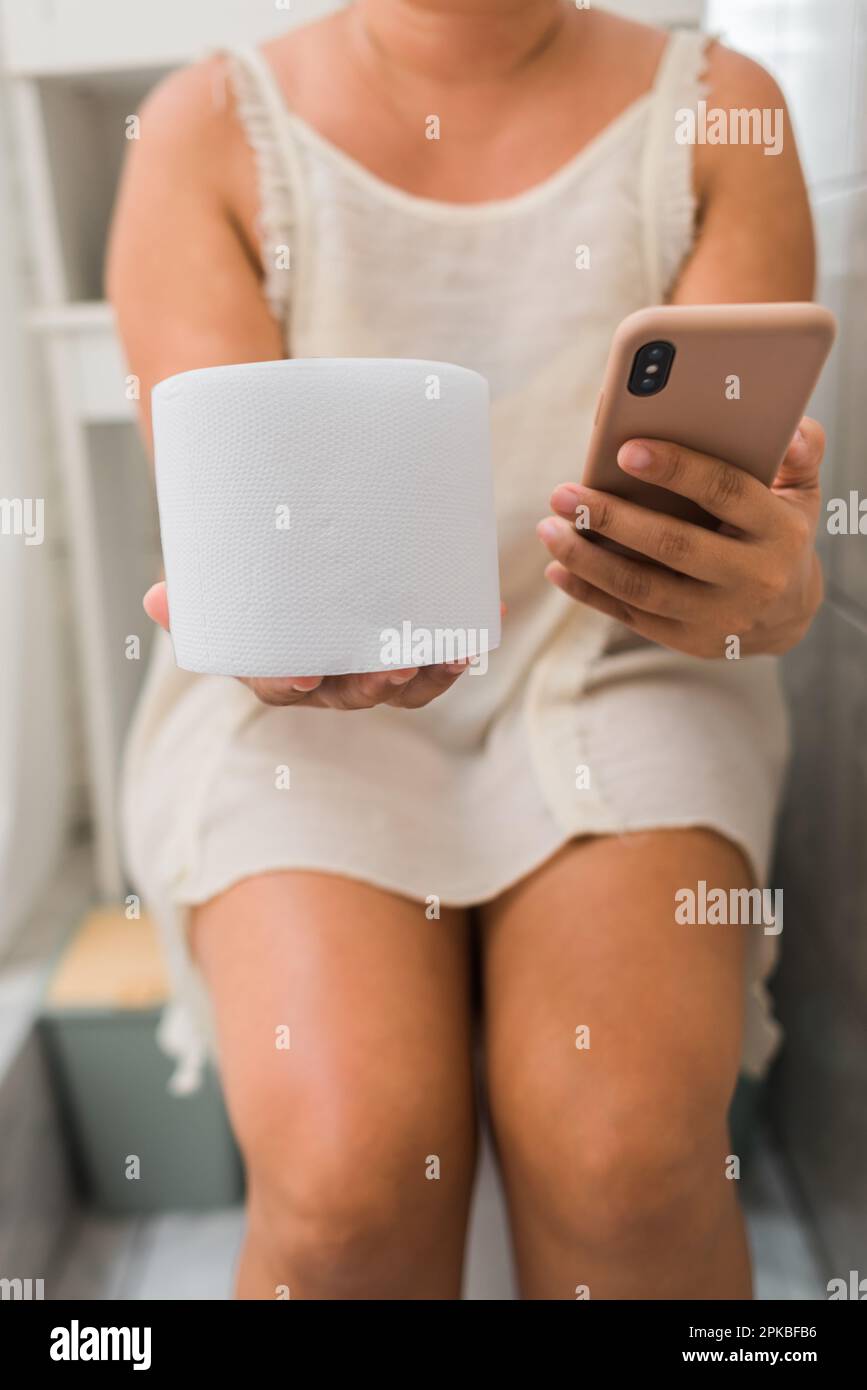 woman holds her cell phone and toilet paper in her hands while sitting on the toilet of her home. Stock Photo